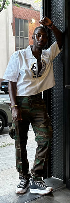 Person wearing a white shirt with a logo and camouflage pants, standing on a sidewalk.
