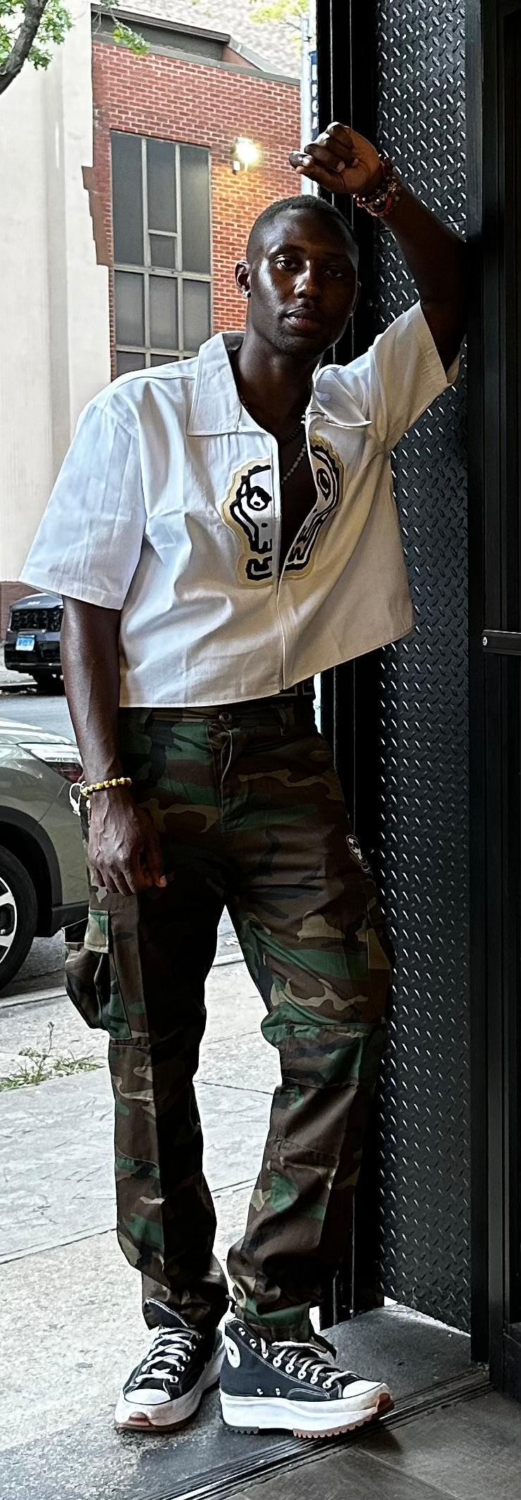 Person wearing a white shirt with a logo and camouflage pants, standing on a sidewalk.